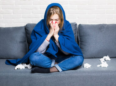 Sick woman with rheum and headache holding napkin, sitting on sofa with coverlet and pills at home